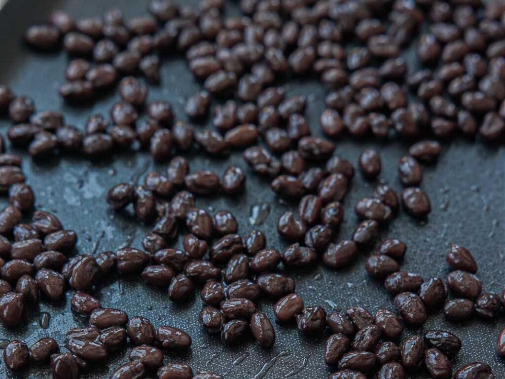 Close-up of black beans spread out on a dark, nonstick surface, glistening with moisture.
